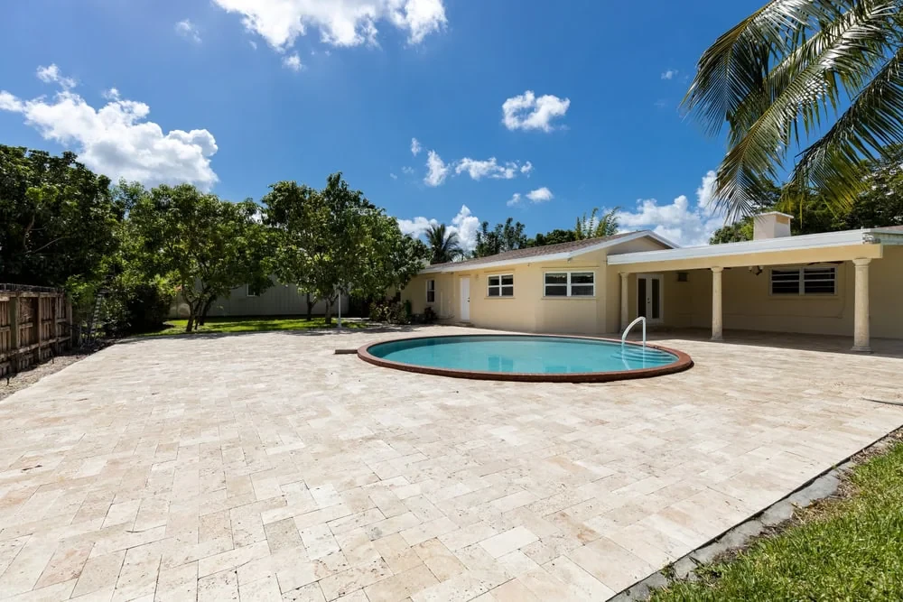 Single-story beige house with a covered patio and round swimming pool in a spacious backyard. Professionally maintained by landscaping services Waco, TX, the area features tiled paving, lush grass, trees, and a wooden fence under a sunny blue sky.