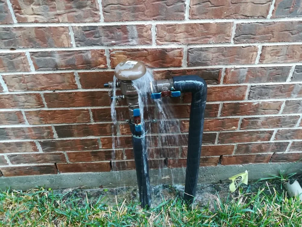 Water is leaking heavily from an outdoor pipe connected to a brick wall, with water cascading down onto the grass below. The pipe, partially wrapped in black insulation, may need attention—consider landscaping services Waco, TX for a solution.