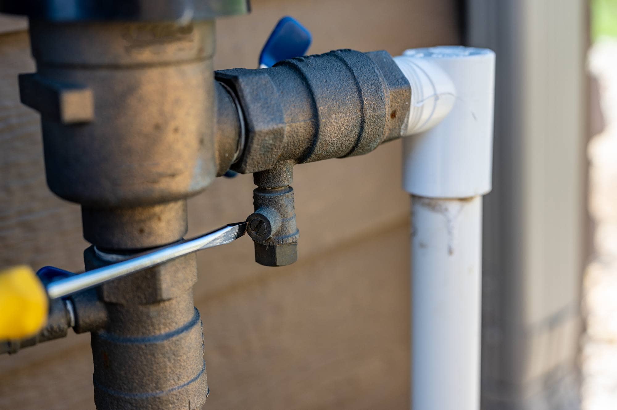A close-up of a screwdriver turning a screw on a metal outdoor plumbing pipe next to a white PVC pipe against a brown wall in Waco, TX—perfect for showcasing expert landscaping services Waco residents trust.
