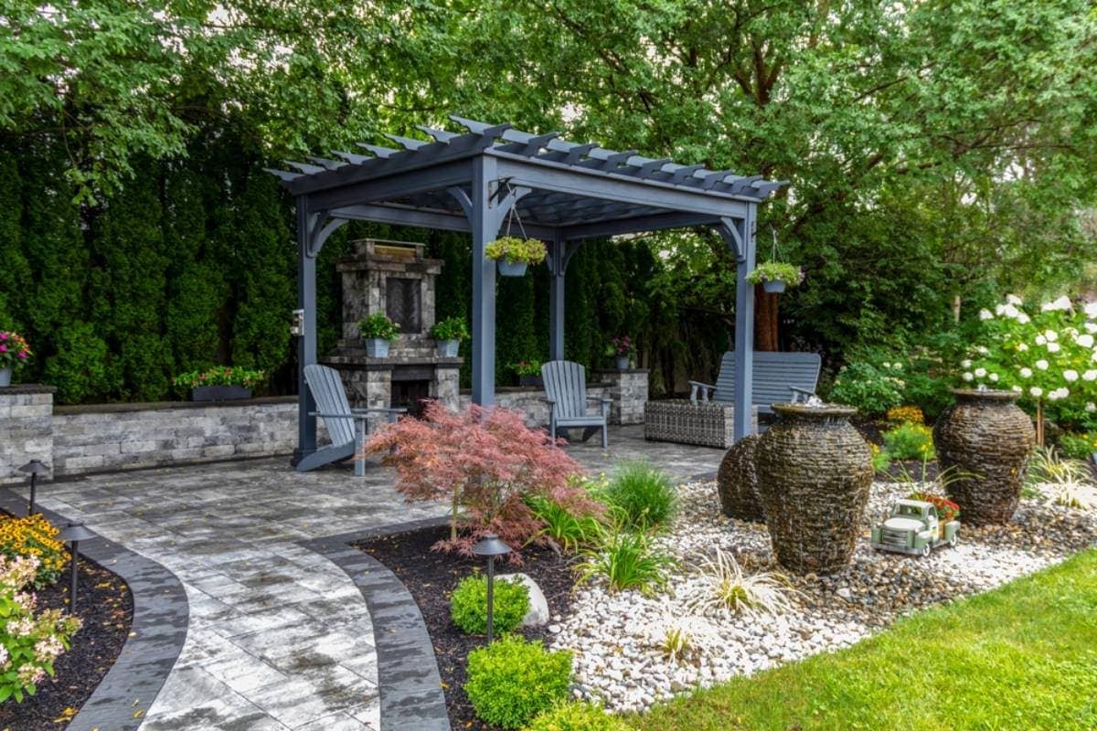 A landscaped backyard with a gray pergola, Adirondack chairs, potted plants, large decorative urn fountains, a stone pathway, and lush greenery—expertly crafted by professional landscaping services in Waco, TX.